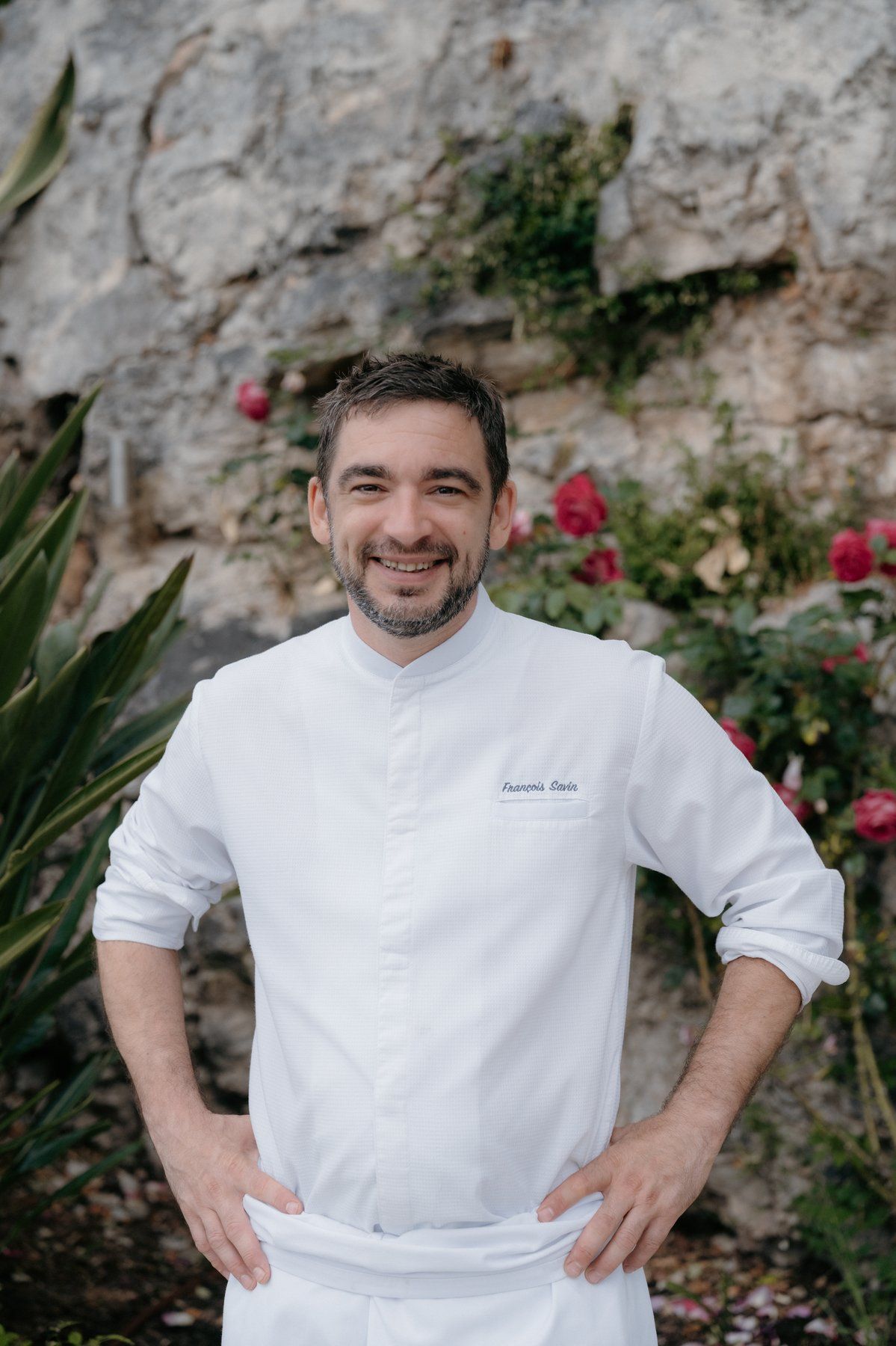 Chef François Savin in his whites, leaning against a stone wall framed by climbing roses.