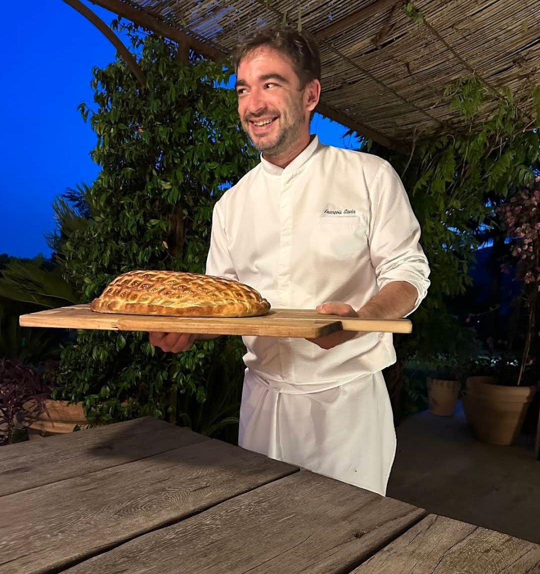 Chef François Savin smiling under a reed pergola, holding a signature beef Wellington on a wooden board at evening light — a private dinner moment.