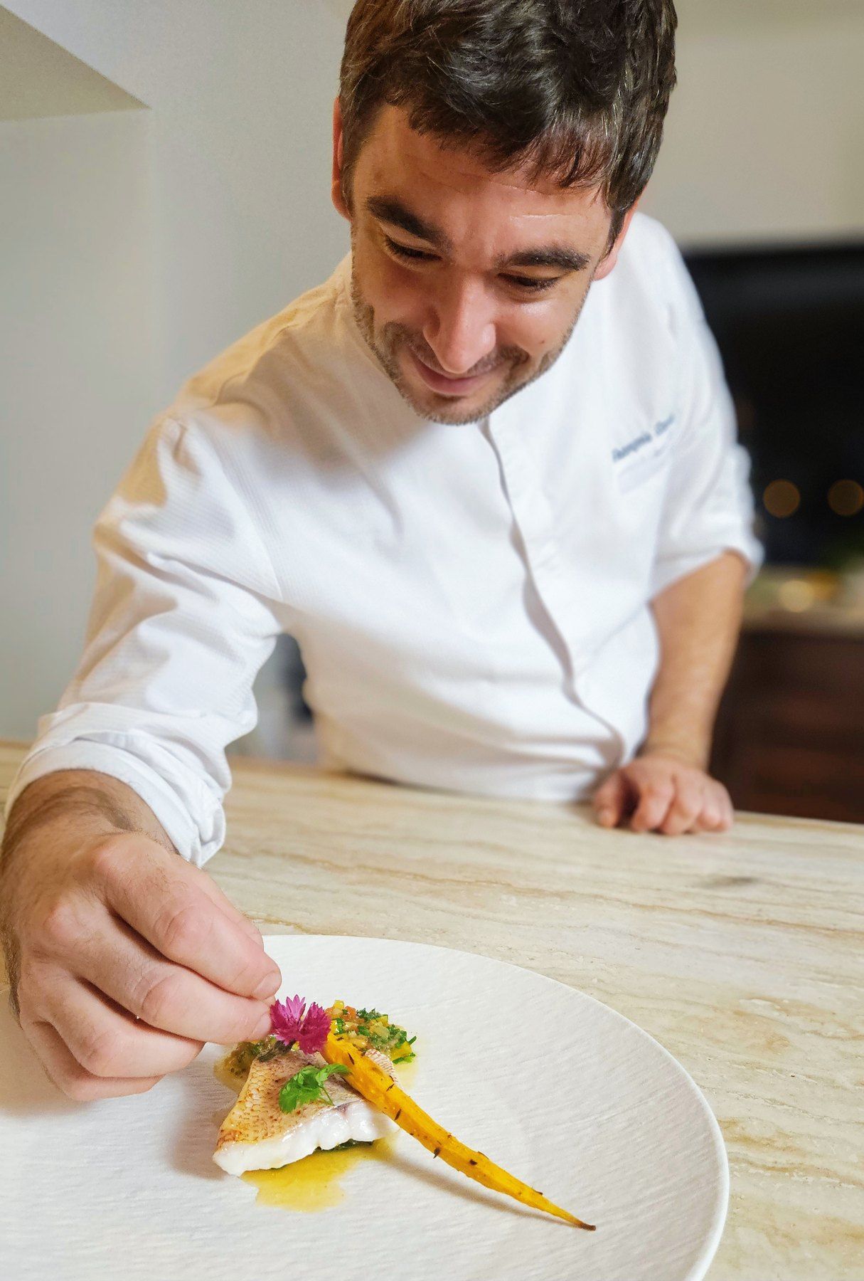 Chef François Savin in his whites, placing an edible flower on a plated red mullet fillet with a glazed carrot.