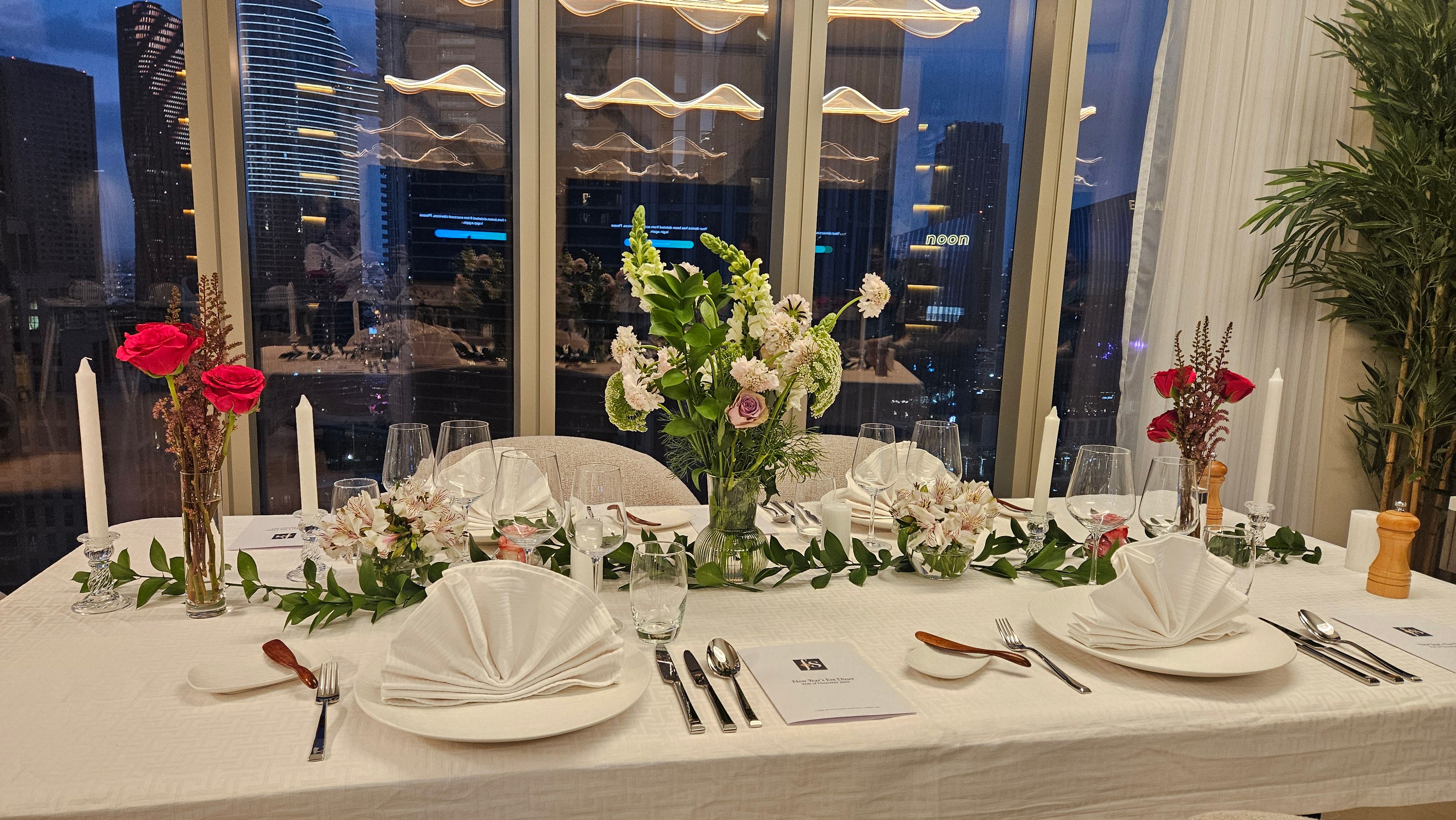 Private dining table dressed for an evening service in a Downtown Dubai residence, the city skyline visible by night through the floor-to-ceiling window.