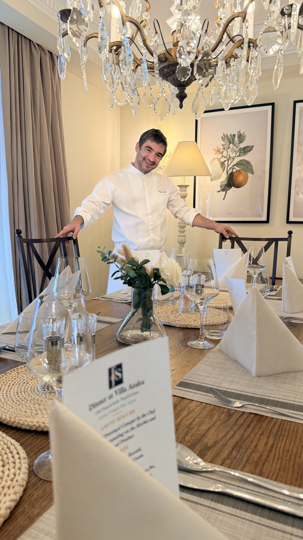 Chef François Savin at a dressed table under a crystal chandelier, printed menus at each place.