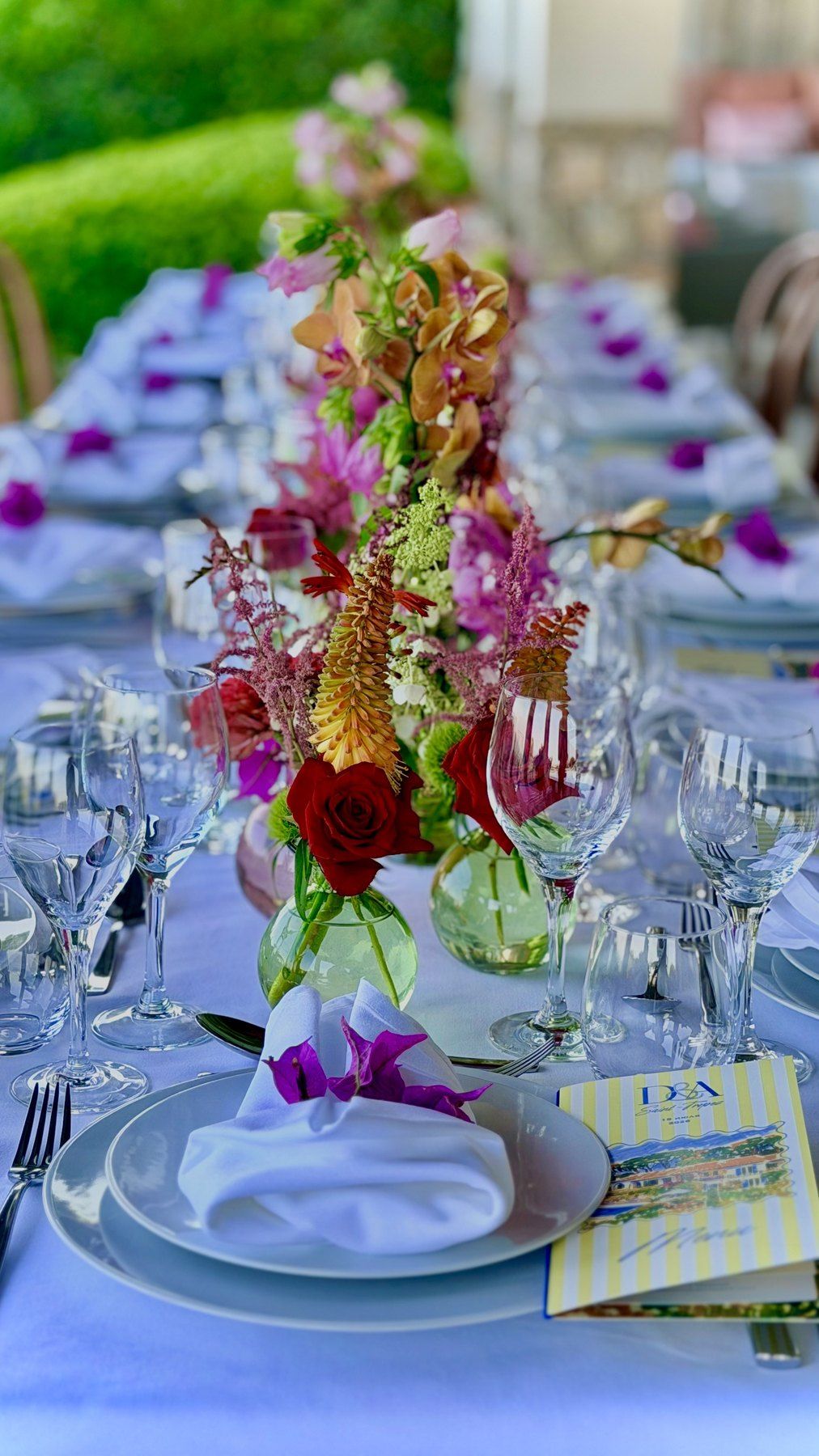 Long table dressed outdoors with a tall floral centrepiece, glassware and folded napkins.