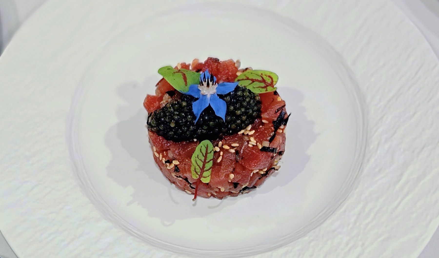 Red tuna tartare topped with caviar, a borage flower and red-veined sorrel leaves, on a white plate.