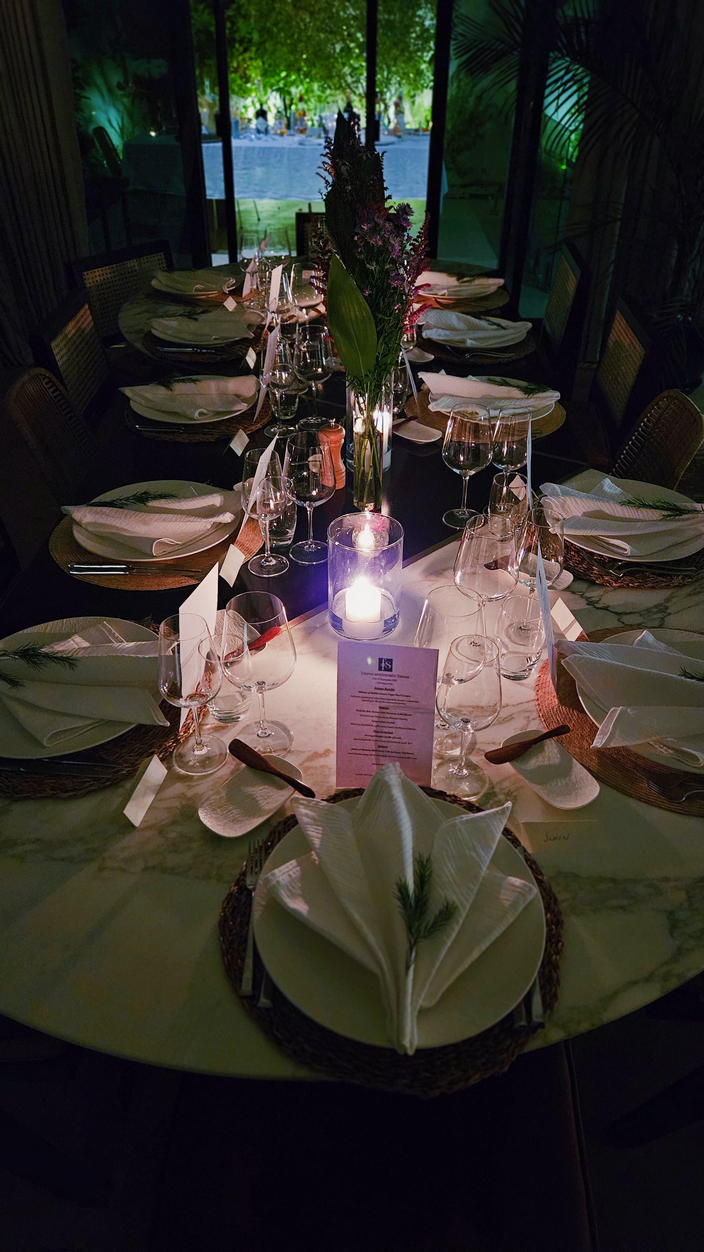 Long villa-banquet table dressed for an evening service on Palm Jumeirah, the swimming pool and palm trees lit beyond the open glass doors.