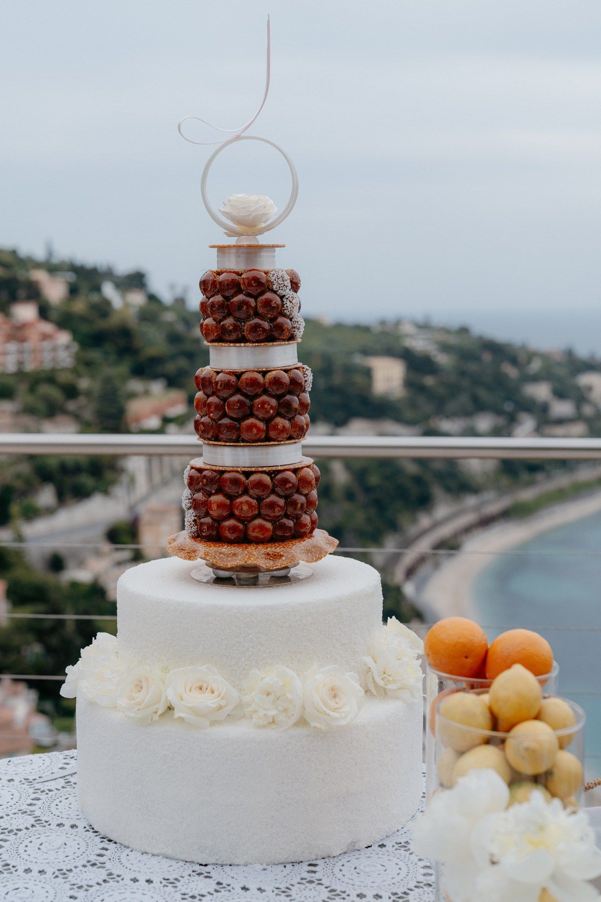 Tiered wedding cake decorated with citrus and peaches, sea view in the background.