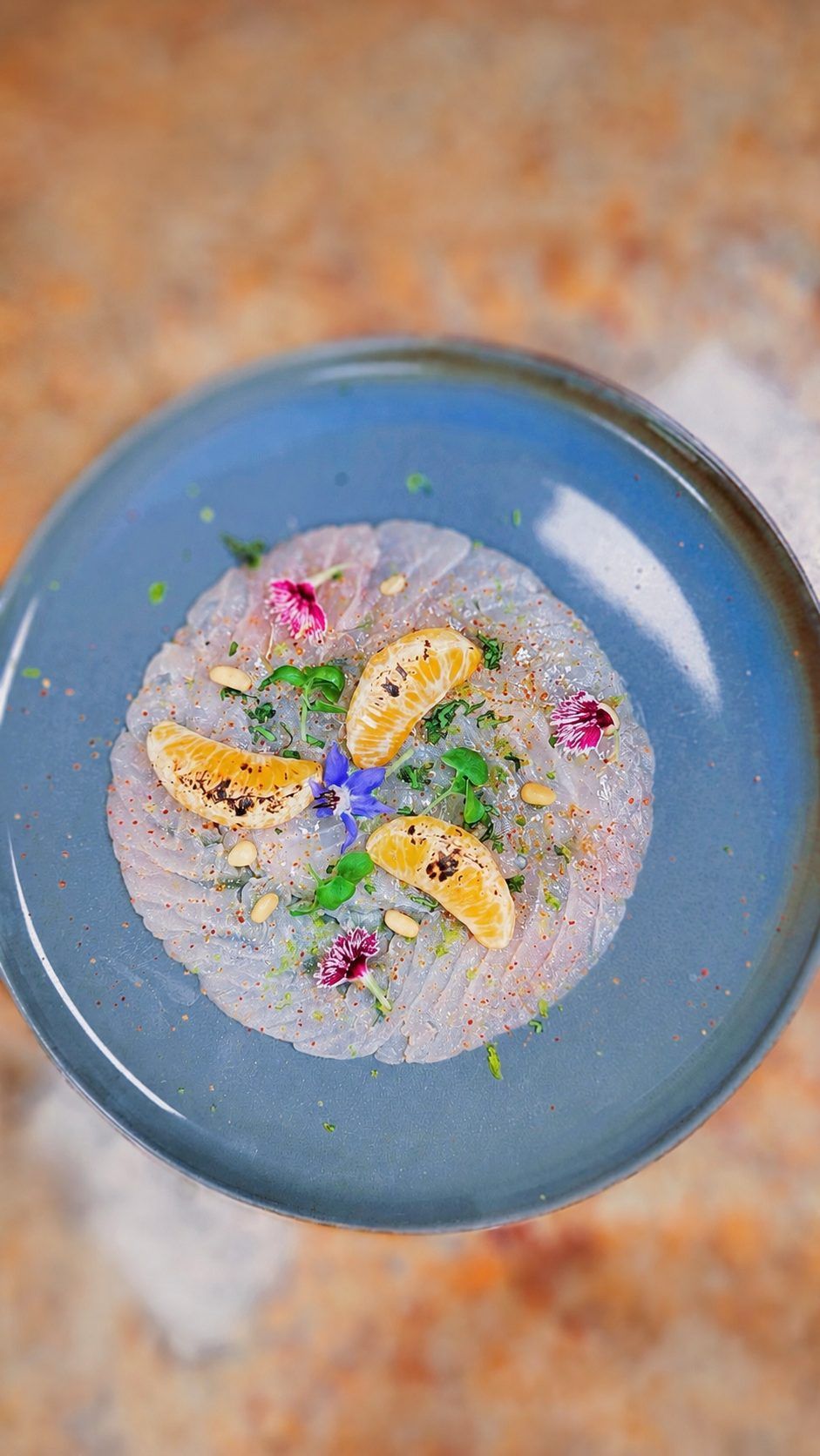 Fish carpaccio with citrus and edible flowers on a blue plate.