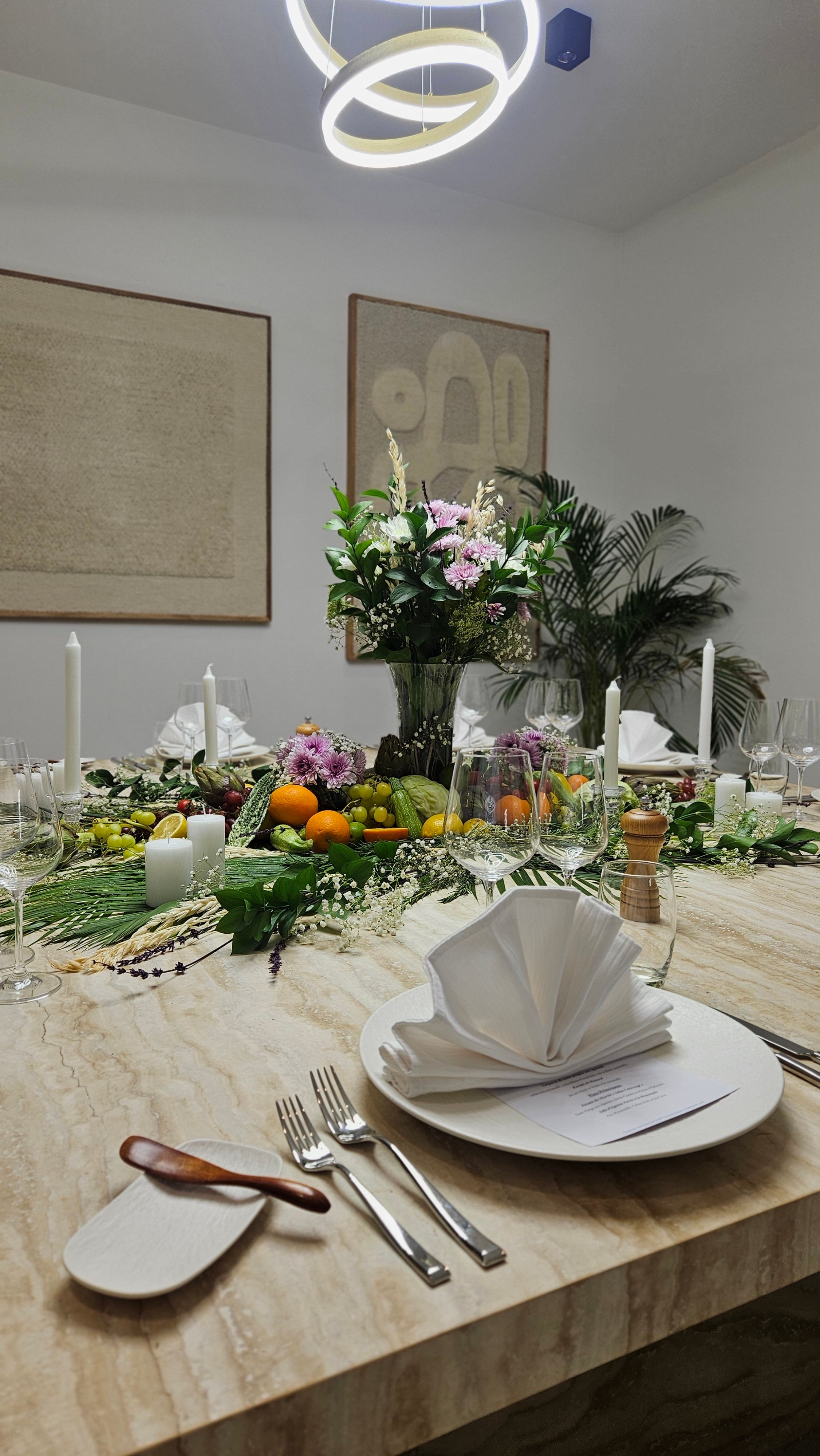 Long private-dining table in a contemporary Dubai residence, dressed with a centerpiece of fresh citrus, grapes, herbs and pale florals, candelabras and folded napkins set for the evening.