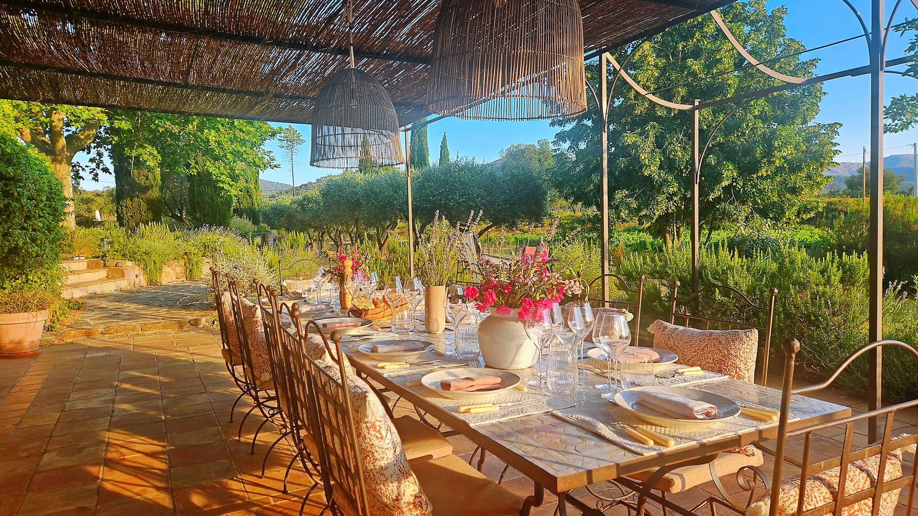 Long villa-terrace table dressed under bamboo shades, garden beyond.